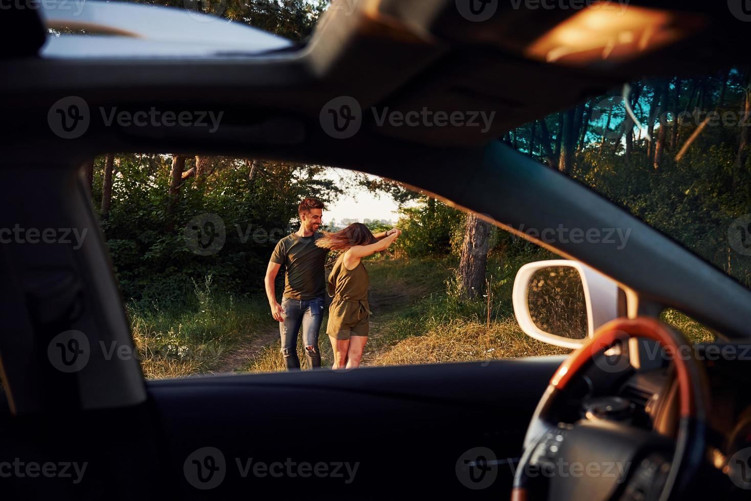 View from the car's interior. Steering wheel, side mirror. Beautiful young couple have a good time in the forest at daytime photo