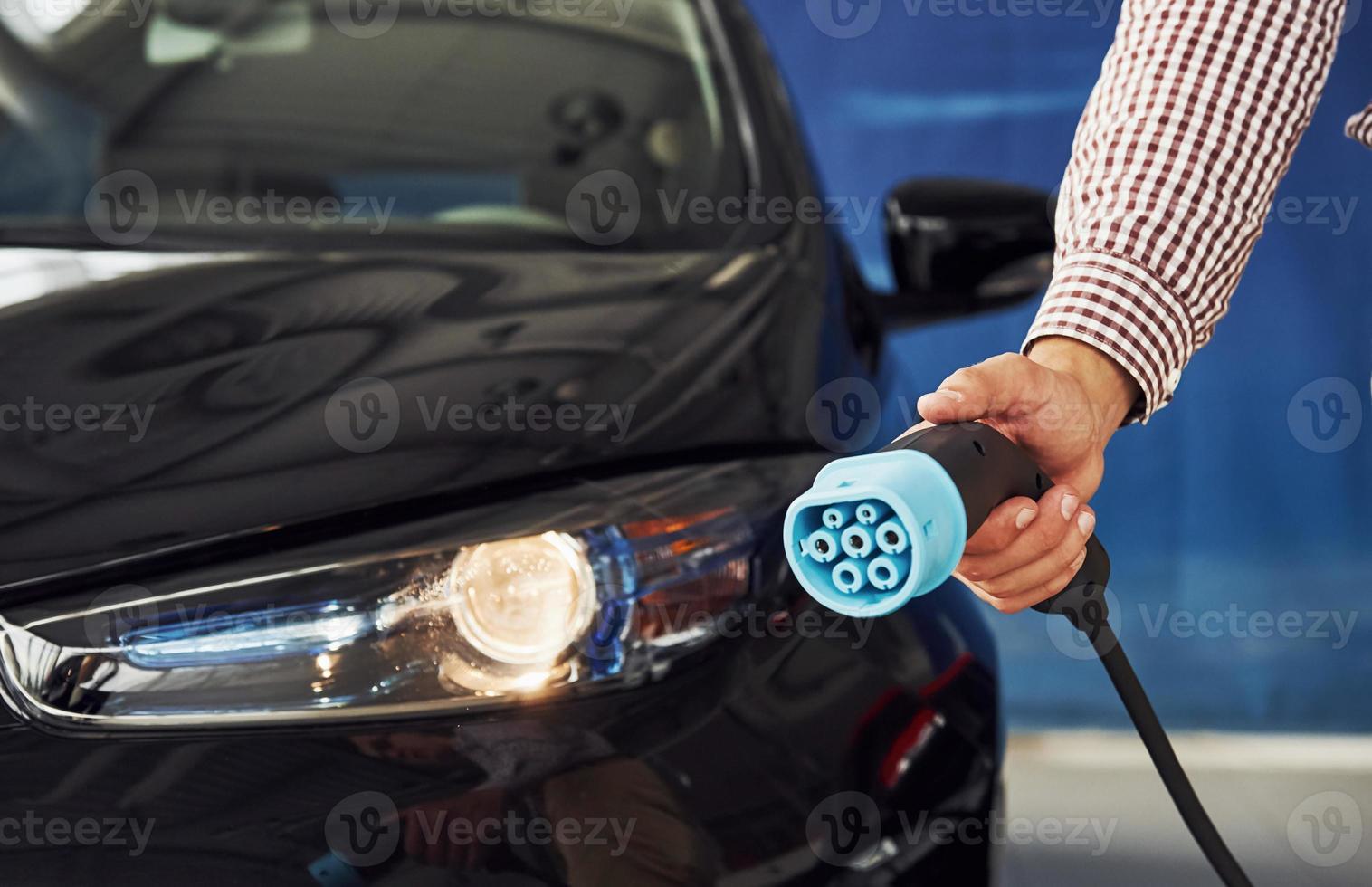 Man stands against an electric car and holds the charging cable photo