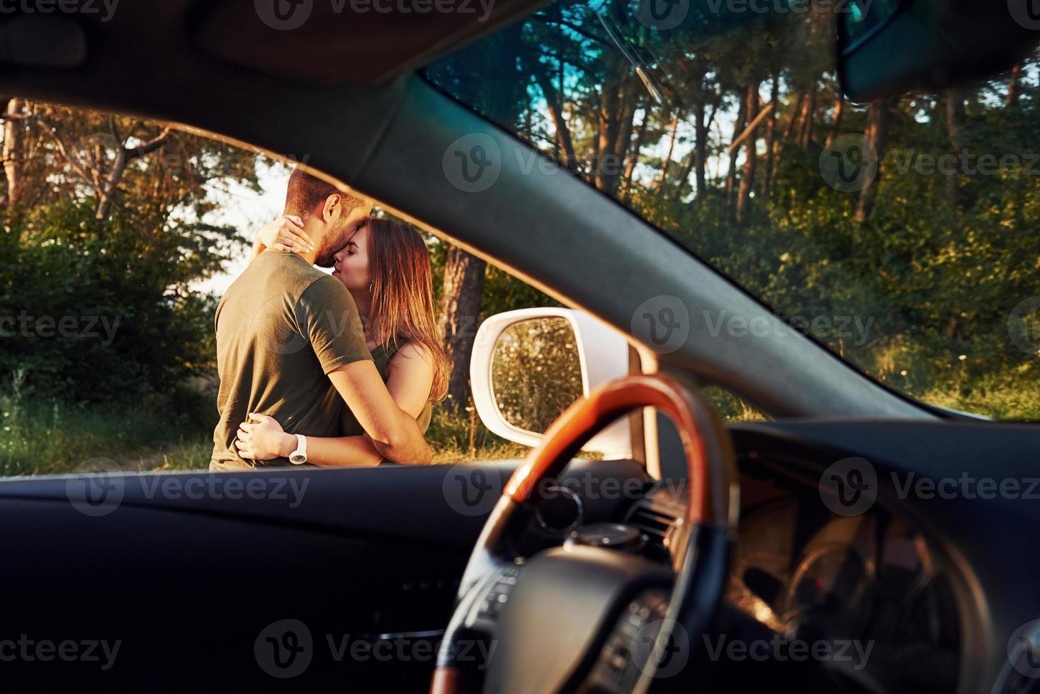 View from the car's interior. Steering wheel, side mirror. Beautiful young couple have a good time in the forest at daytime photo