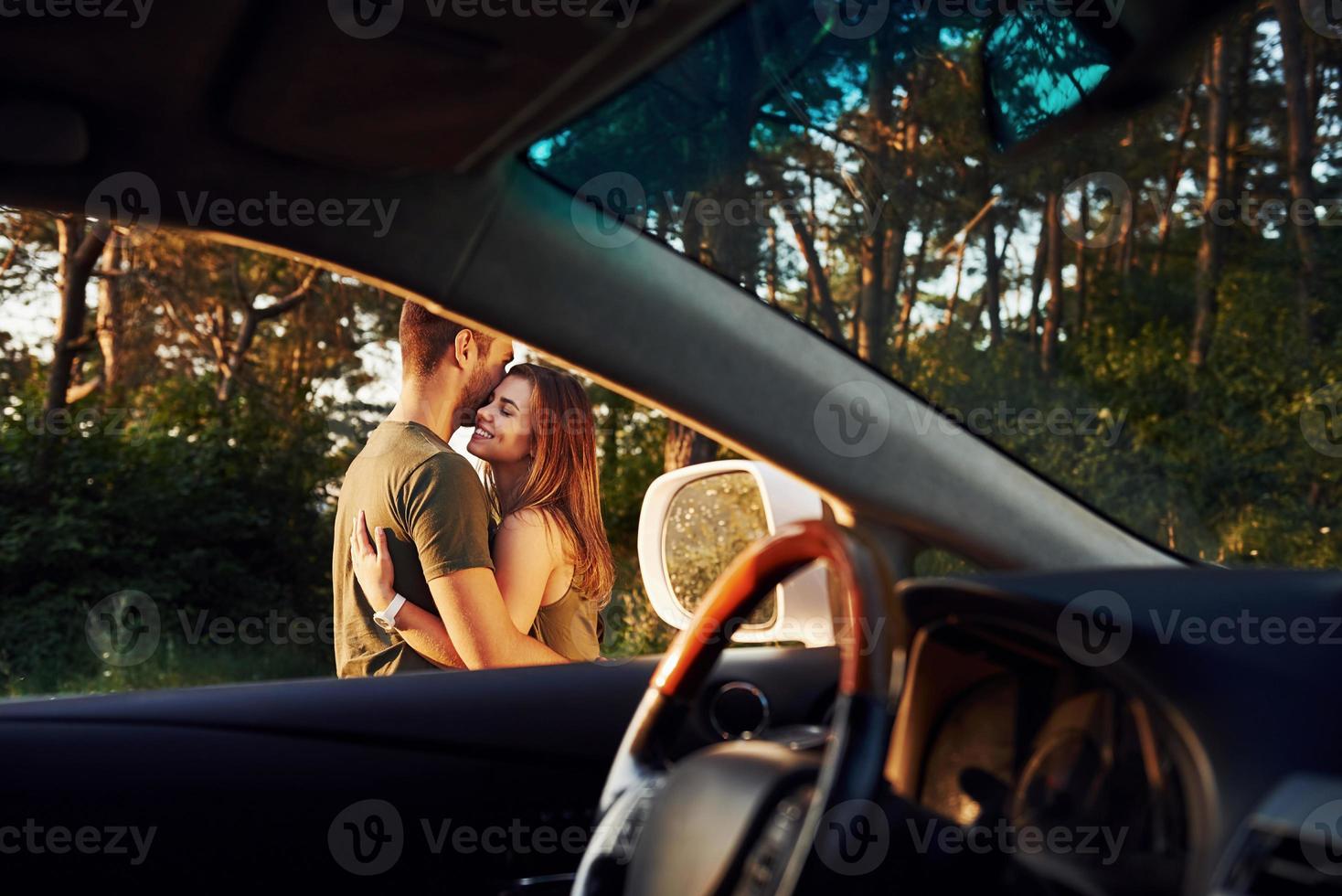 View from the car's interior. Steering wheel, side mirror. Beautiful young couple have a good time in the forest at daytime photo