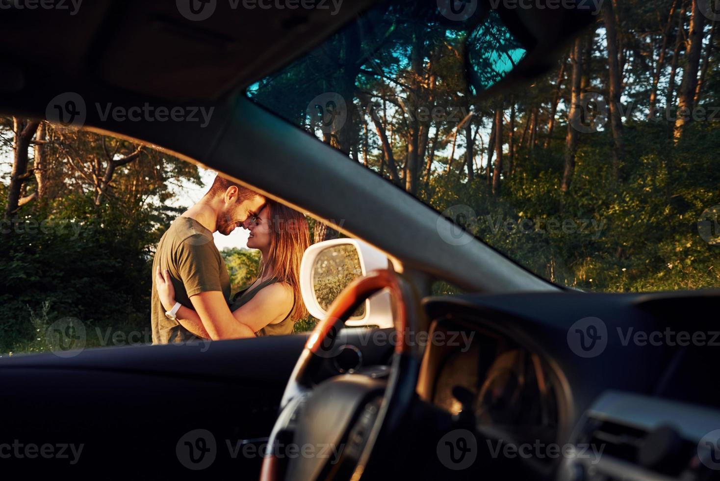 View from the car's interior. Steering wheel, side mirror. Beautiful young couple have a good time in the forest at daytime photo