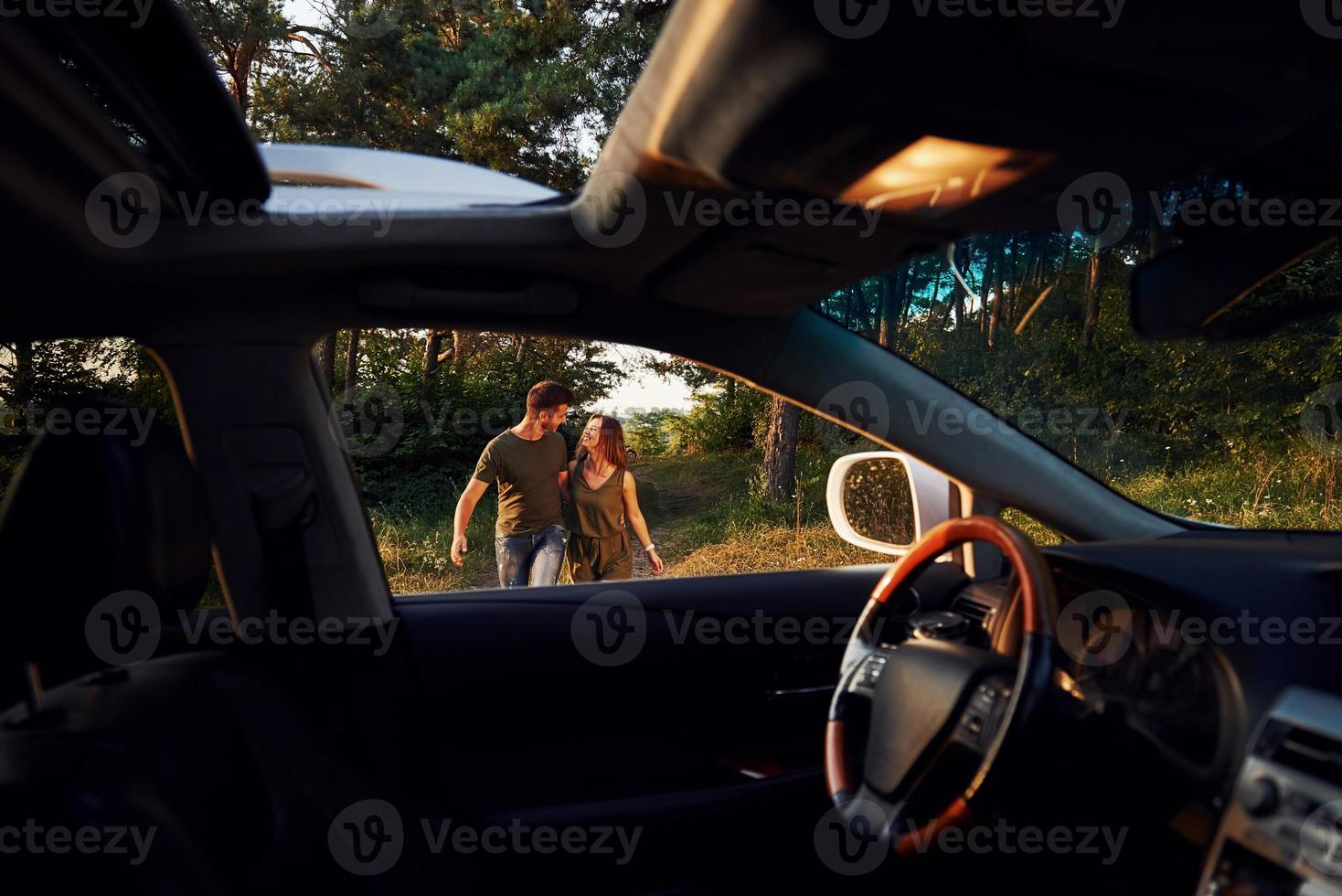 View from the car's interior. Steering wheel, side mirror. Beautiful young couple have a good time in the forest at daytime photo