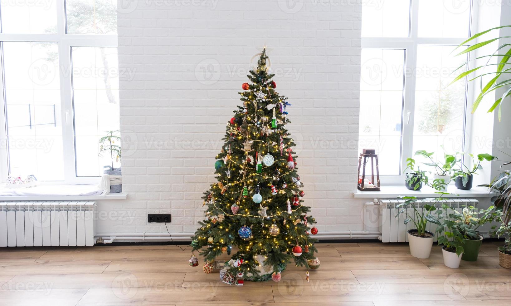 Christmas tree in the white interior of a house with large windows. Glowing fairy lights garlands interior decoration of the studio room. Potted plants in the home photo