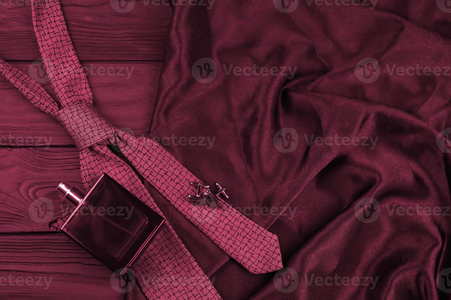 A bottle of mens cologne and cufflinks with blue tie lie on a black luxury fabric background on a wooden table. Mens accessories Image toned in Viva Magenta, color of the 2023 year photo