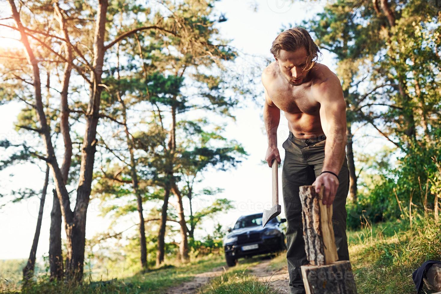 leñador con un hacha cortando madera. un hombre guapo sin camisa con un tipo de cuerpo musculoso está en el bosque durante el día foto