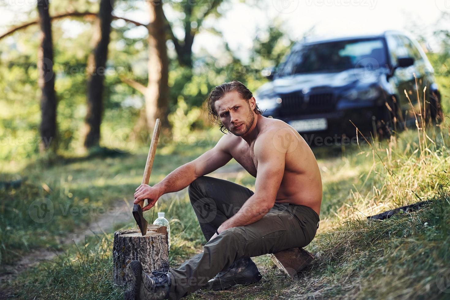 Holds wooden axe. Handsome shirtless man with muscular body type is in the forest at daytime photo