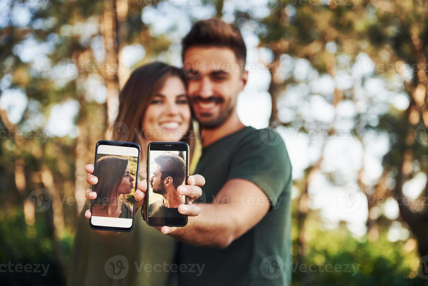 sosteniendo dos teléfonos inteligentes con fotos de ellos. hermosa pareja joven diviértete en el bosque durante el día