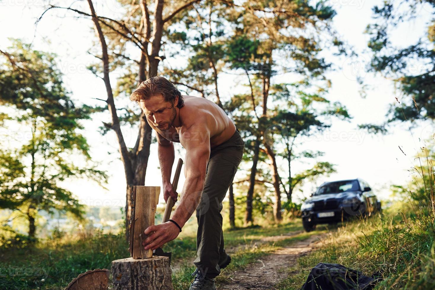 Woodsman with an axe cutting wood. Handsome shirtless man with muscular body type is in the forest at daytime photo