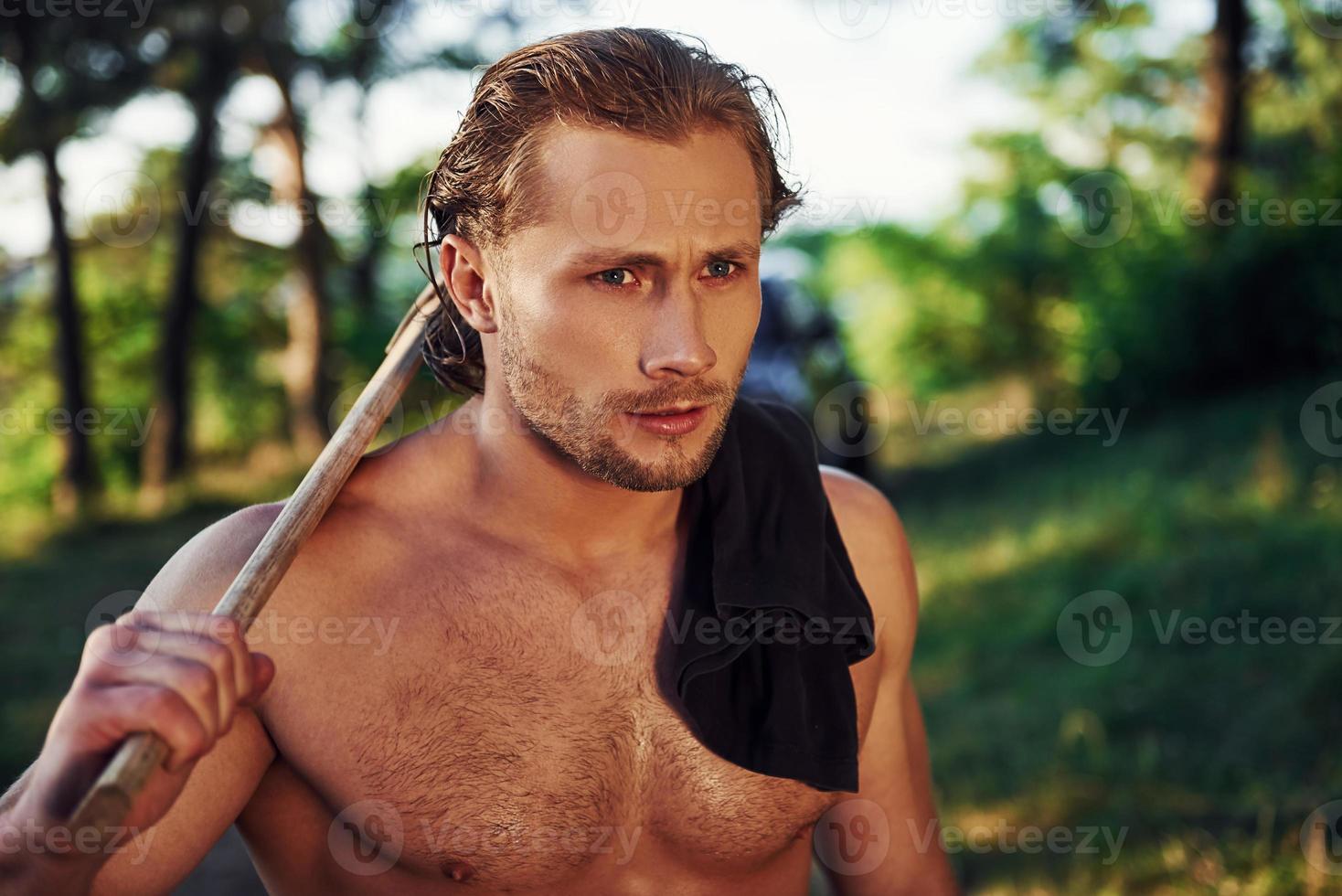 Close up portrait of woodsman with axe in hand. Handsome shirtless man with muscular body type is in the forest at daytime photo