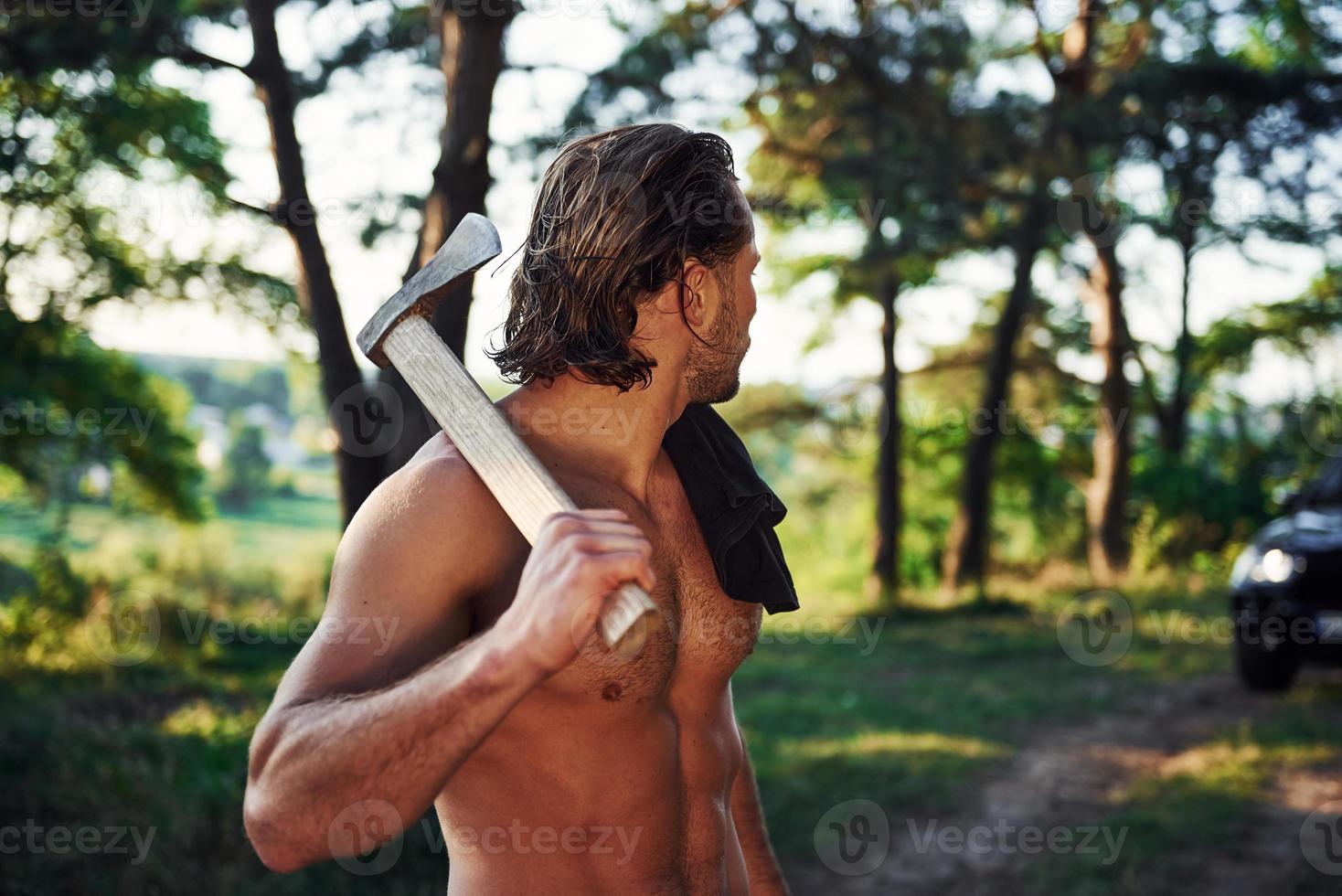 Close up portrait of woodsman with axe in hand. Handsome shirtless man with muscular body type is in the forest at daytime photo