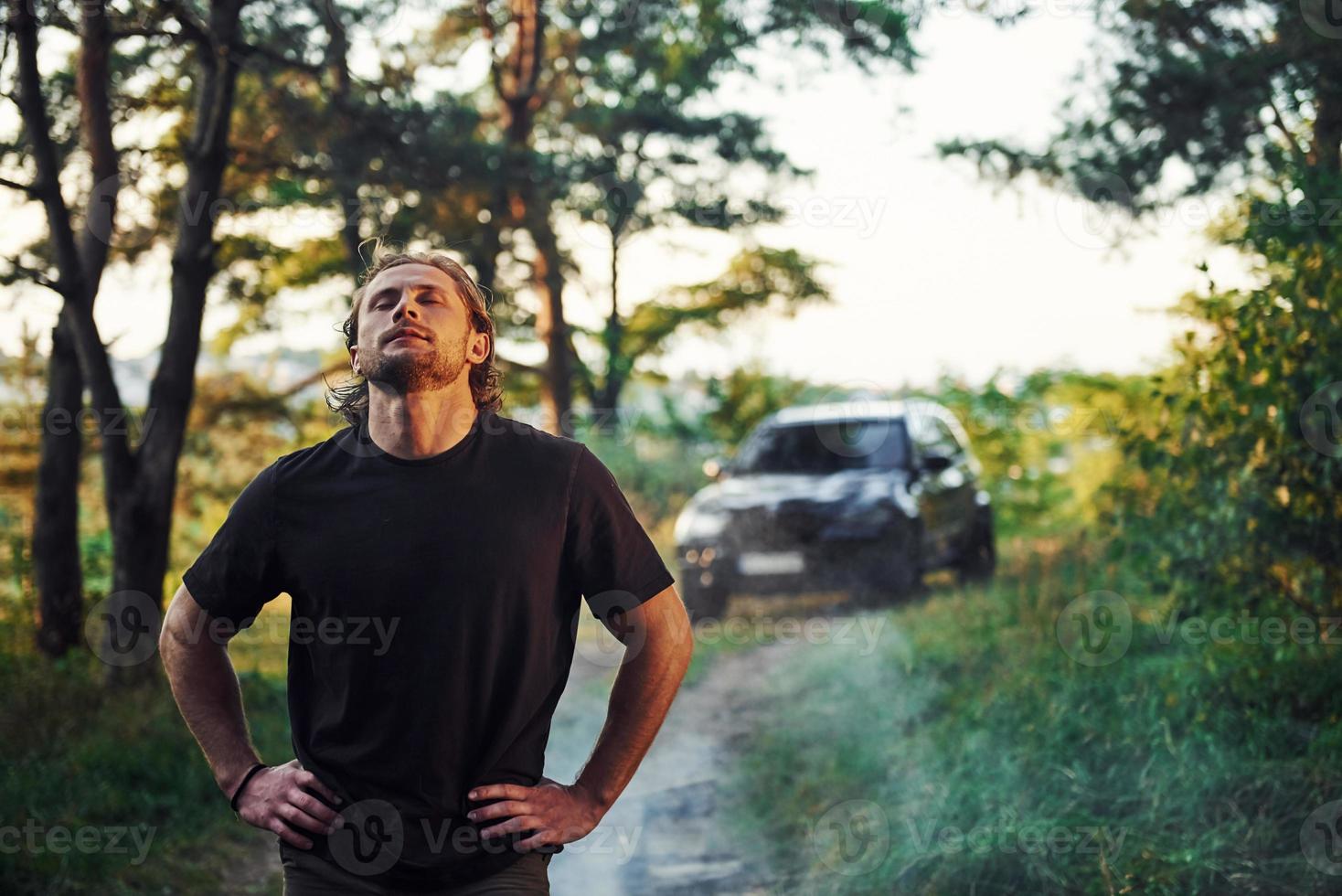 Black automobile behind. Portrait of man that stands in the forest and looks at nature photo