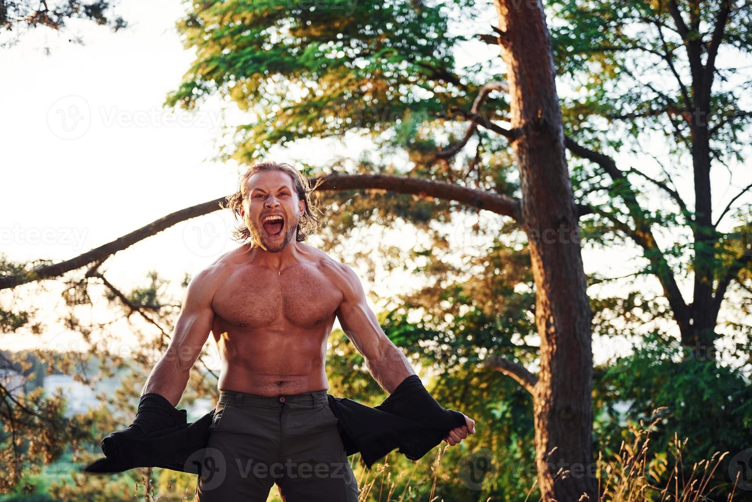 Looks behind. Handsome shirtless man with muscular body type is in the forest at daytime photo