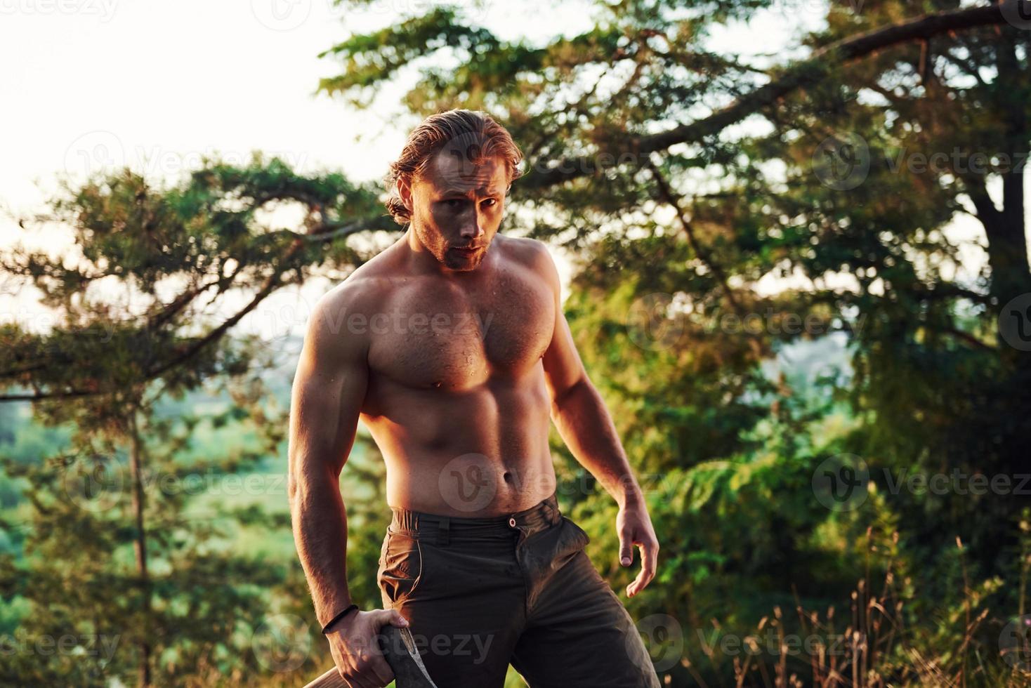 listo para el trabajo. leñador confiado. un hombre guapo sin camisa con un tipo de cuerpo musculoso está en el bosque durante el día foto