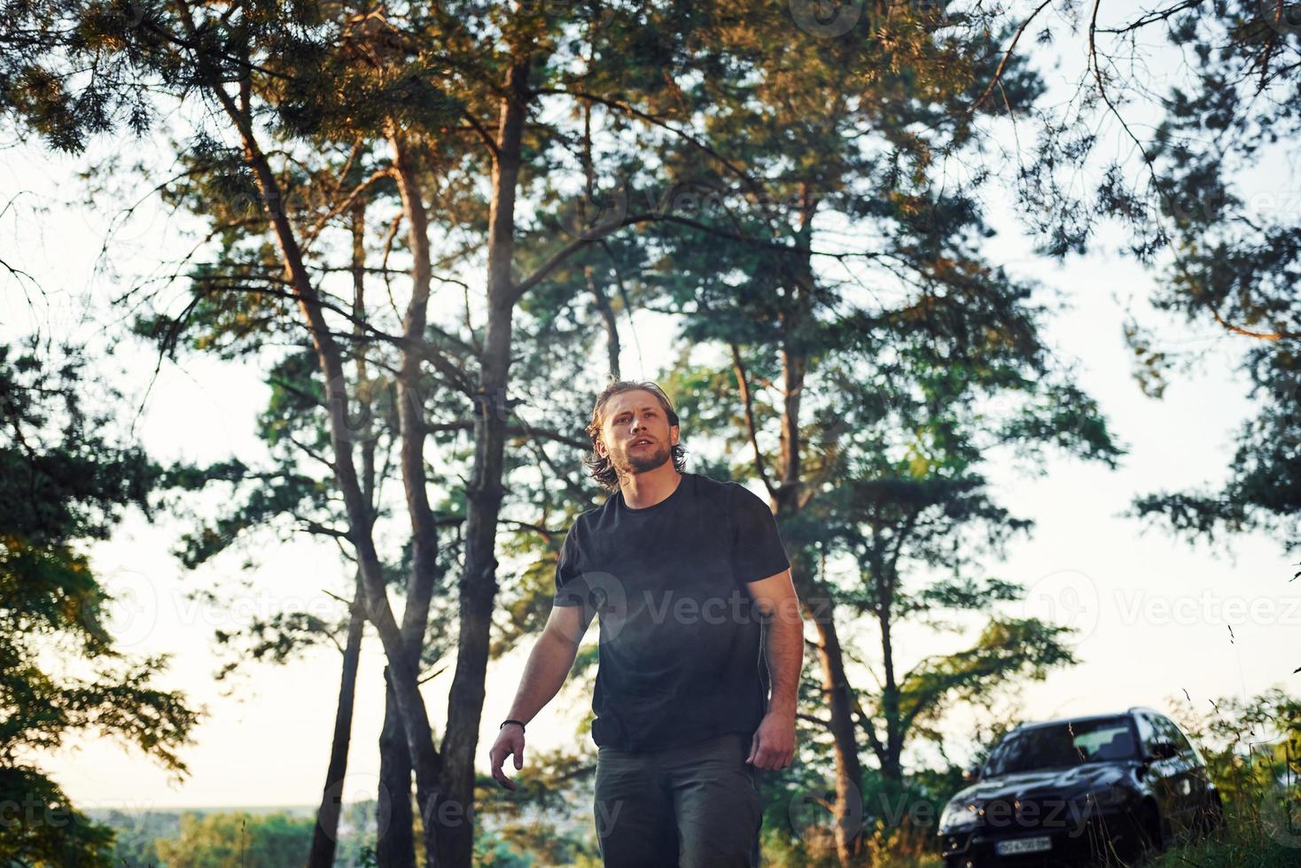 automóvil negro detrás. retrato del hombre que se para en el bosque y mira la naturaleza foto