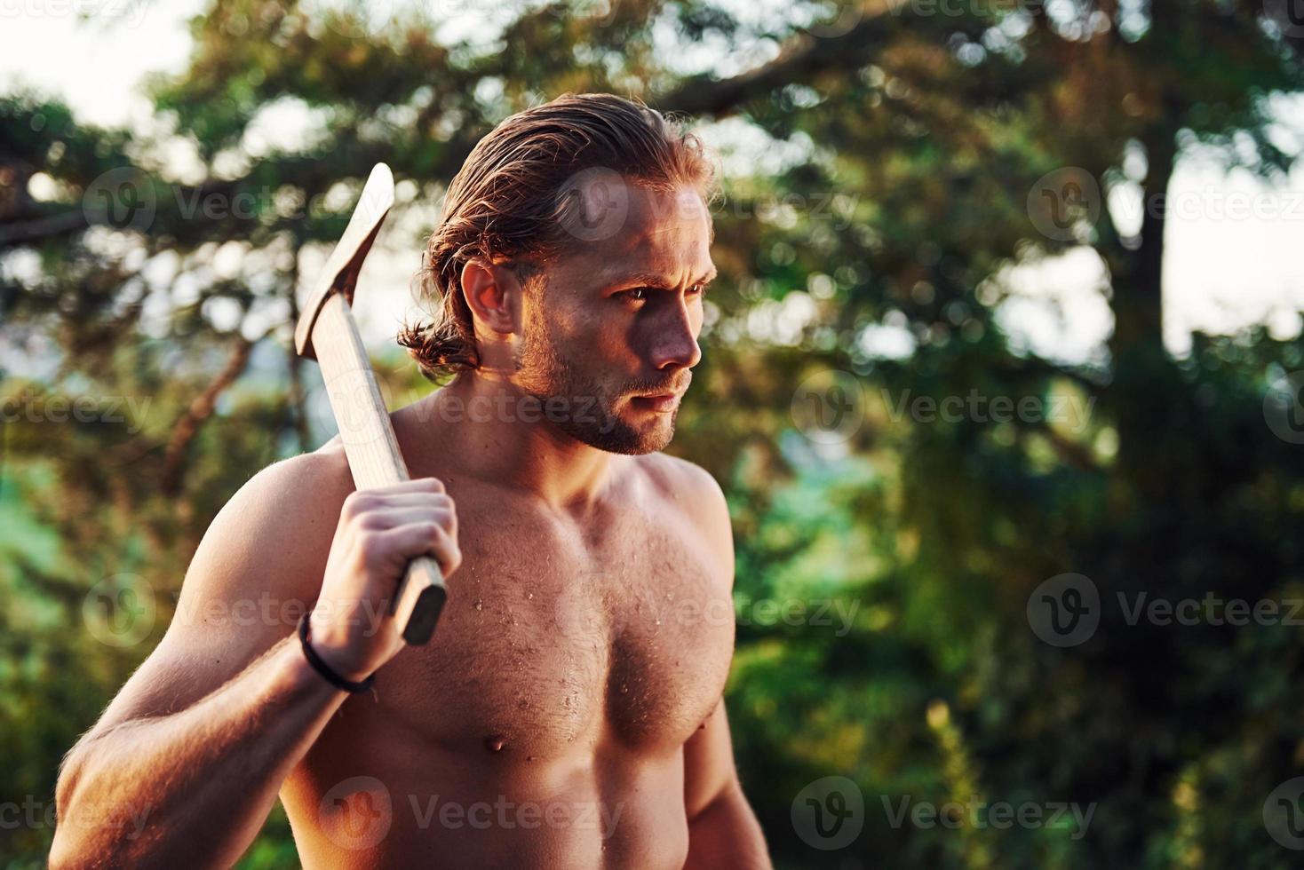 Ready for the work. Confident woodsman. Handsome shirtless man with muscular body type is in the forest at daytime photo