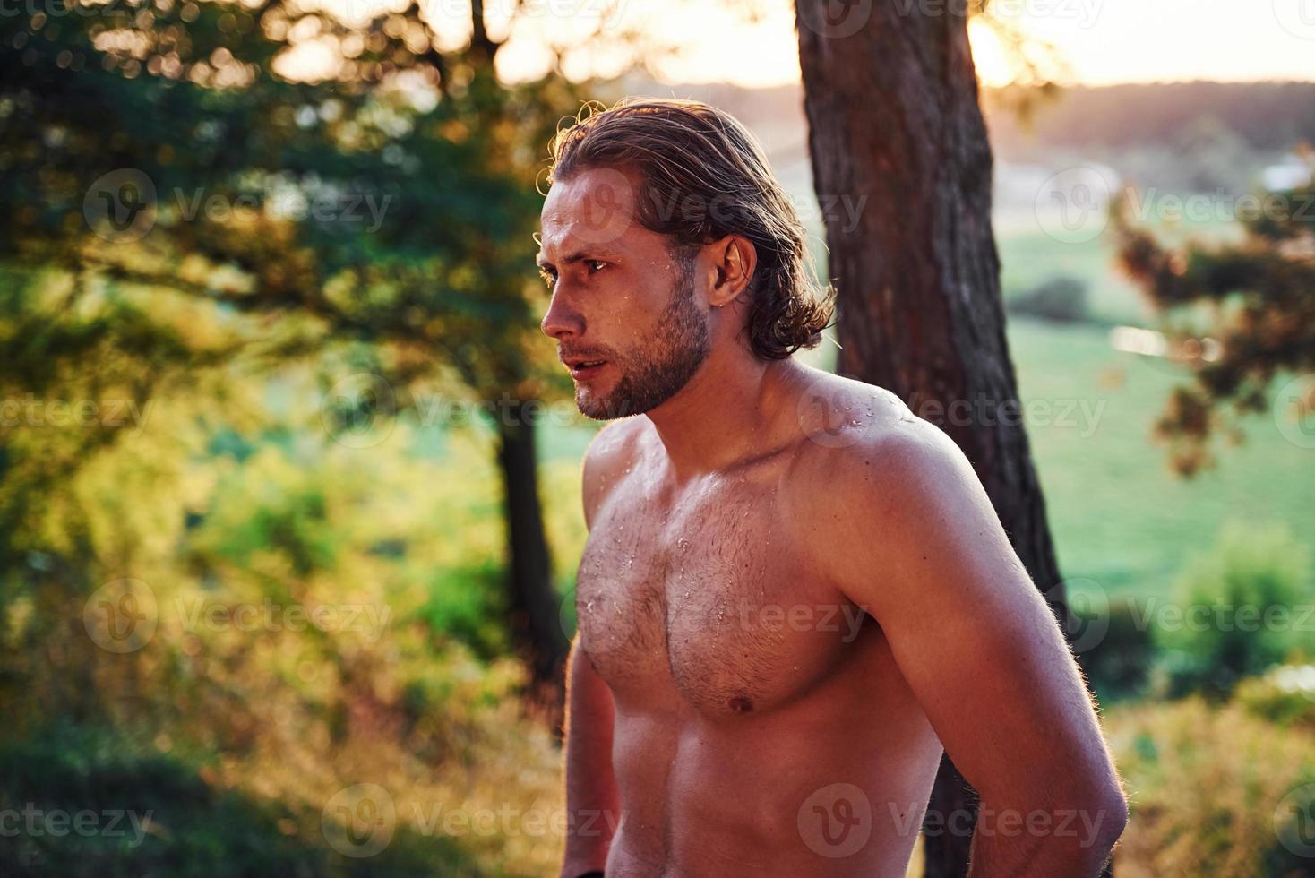 mirada seria y confiada. un hombre guapo sin camisa con un tipo de cuerpo musculoso está en el bosque durante el día foto