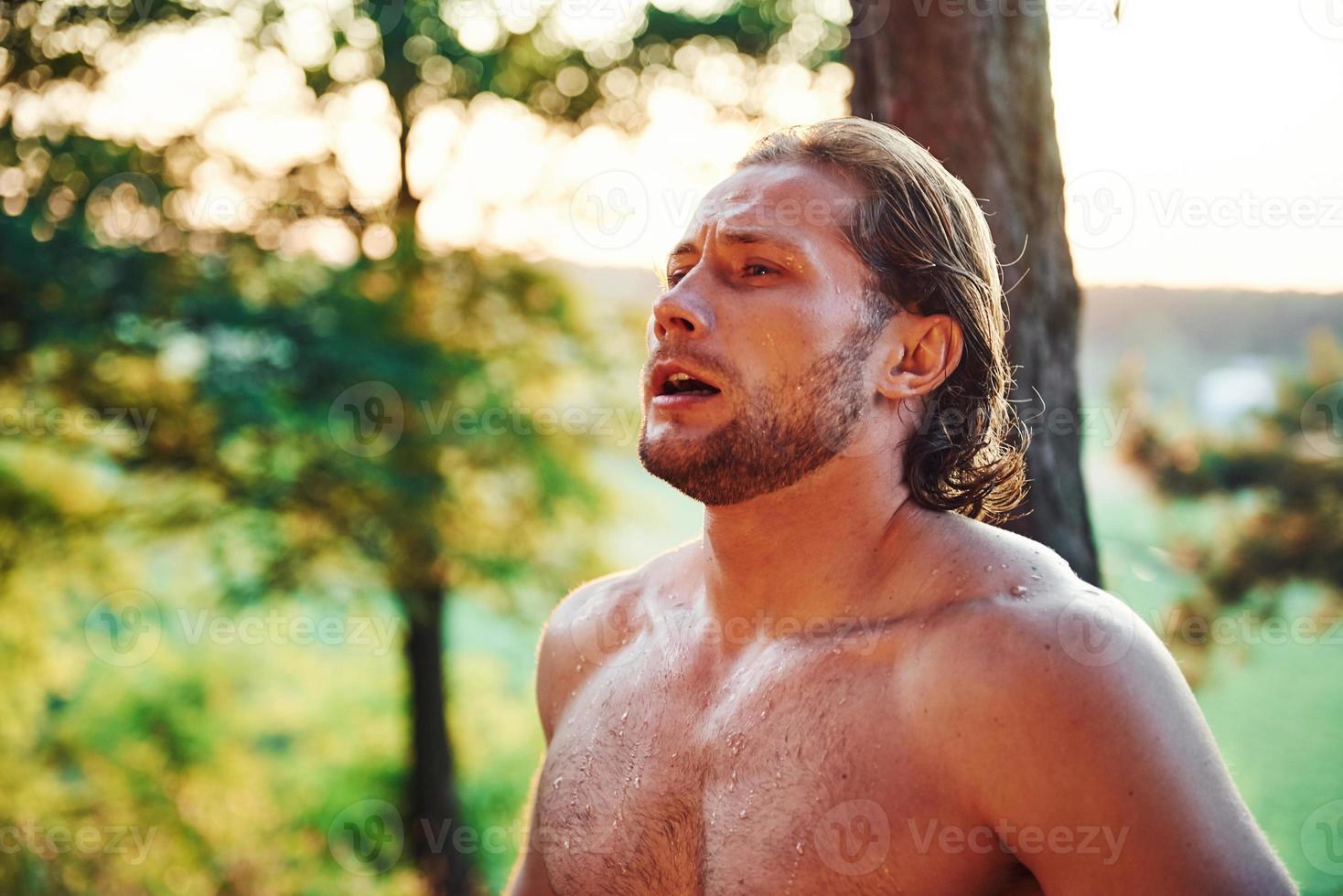 mirada seria y confiada. un hombre guapo sin camisa con un tipo de cuerpo musculoso está en el bosque durante el día foto