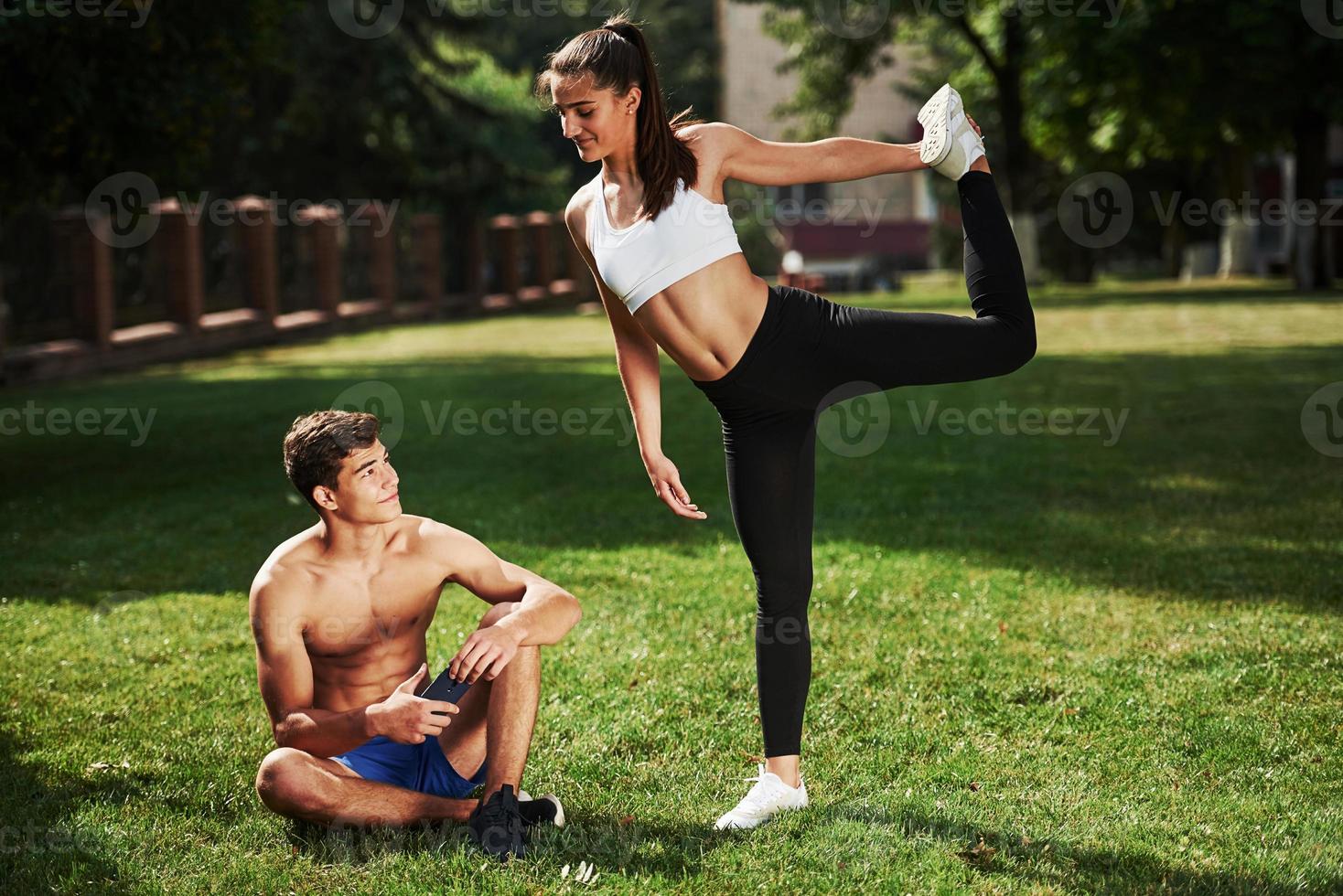 Guy sitting with smartphone in hands and have rest. Man and woman have fitness day in the city at daytime in the park photo