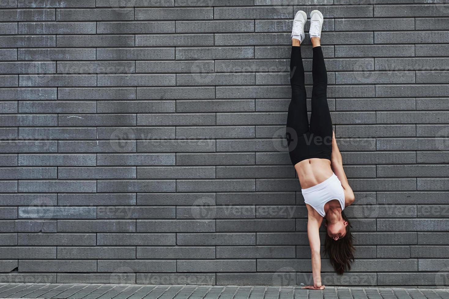 Doing handstand. Young sportive brunette with slim body shape against brick wall in the city at daytime photo