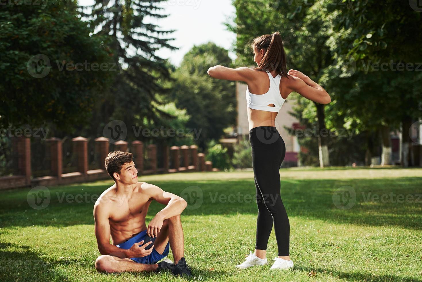 Guy sitting with smartphone in hands and have rest. Man and woman have fitness day in the city at daytime in the park photo