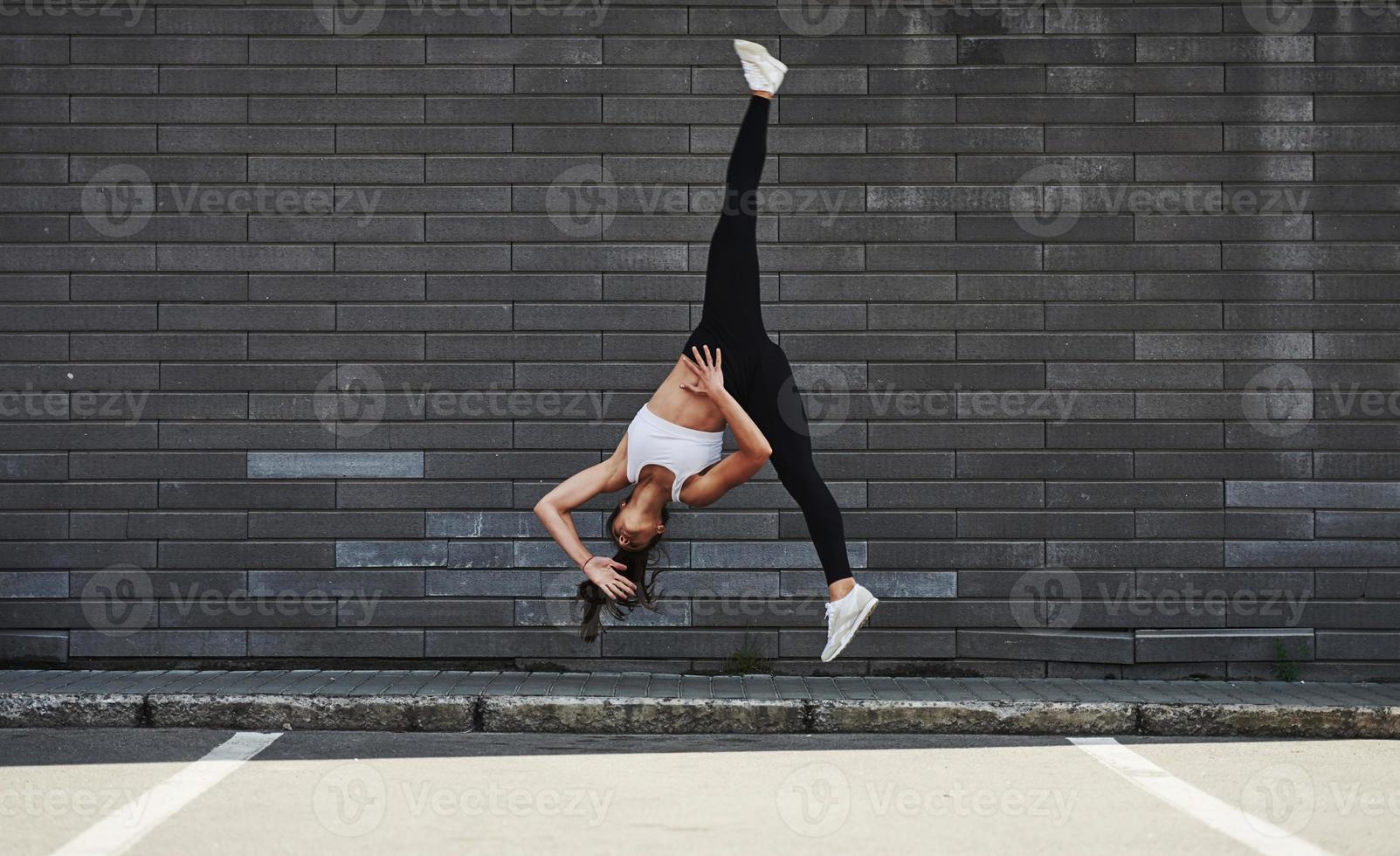 Doing crazy parkour and athletic stunts. Young sportive brunette with slim body shape against brick wall in the city at daytime photo