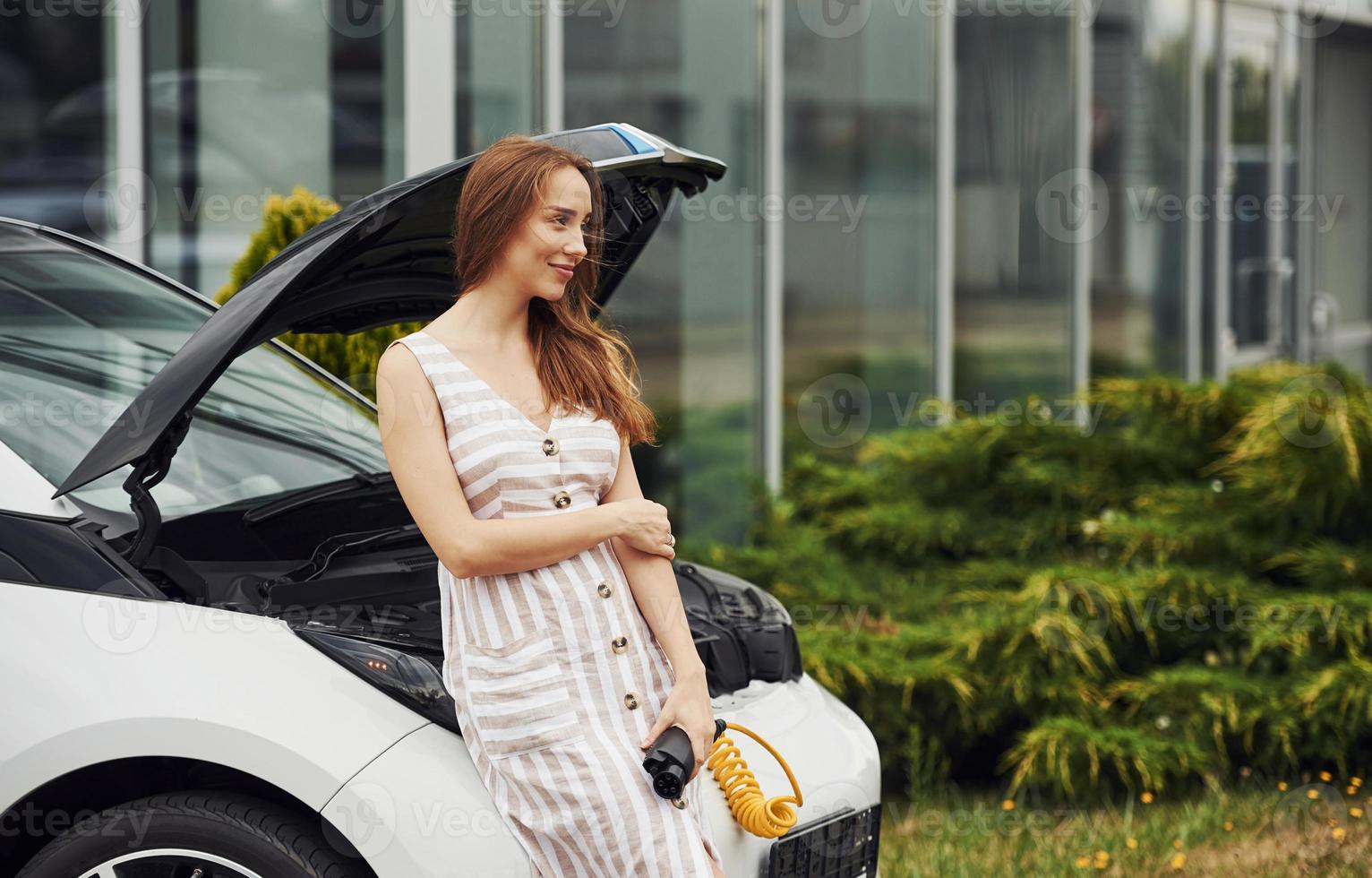Leans on the front of the automobile. Woman on the electric cars charge station at daytime. Brand new vehicle photo