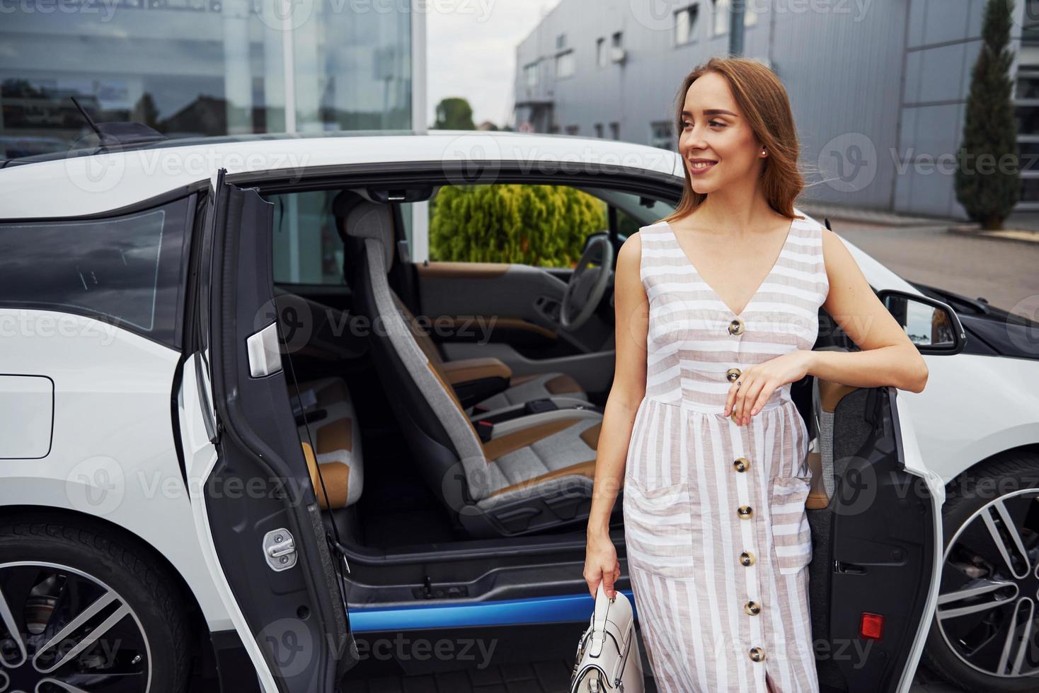 With bag in hand. Woman with blonde hair stands near her white car on the street photo