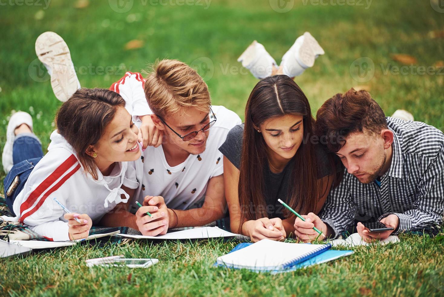 Writing on paper. front view. Group of young students in casual clothes on green grass at daytime photo