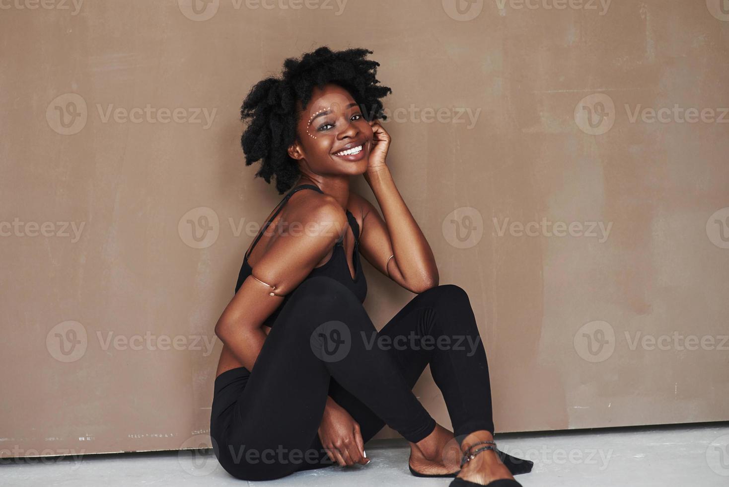 sentado en el suelo. joven y bella mujer afroamericana en el estudio con antecedentes marrones foto