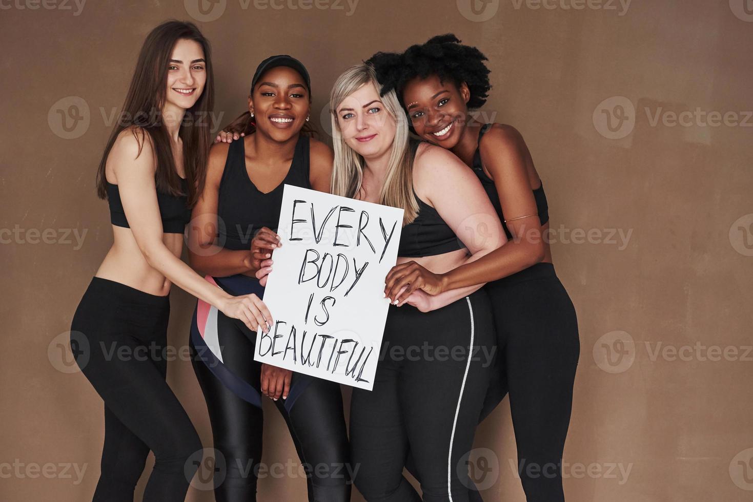 Holding sign with motivational letters. Group of multi ethnic women standing in the studio against brown background photo