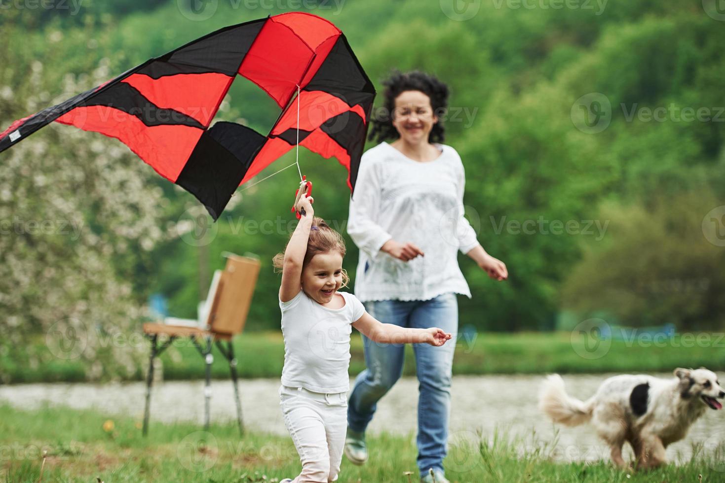 Wonderful dog on right side. Positive female child and grandmother running with red and black colored kite in hands outdoors photo