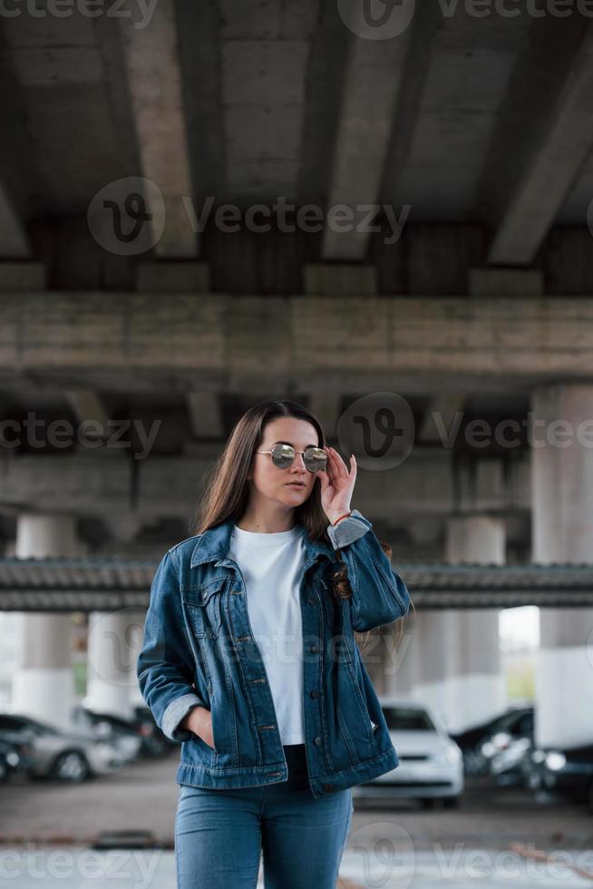 In jeans clothes. Portrait of beautiful young woman standing under the bridge outdoors photo