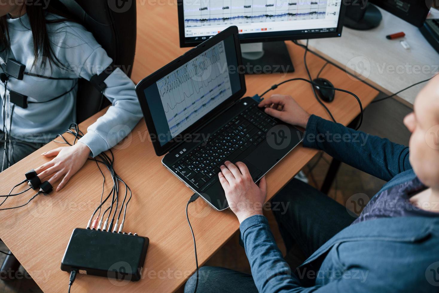 Business competitor is in the chair. Girl passes lie detector in the office. Asking questions. Polygraph test photo