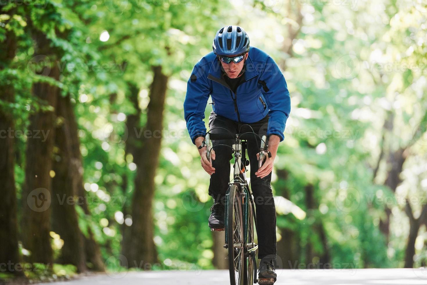 In blue jacket. Cyclist on a bike is on the asphalt road in the forest at sunny day photo