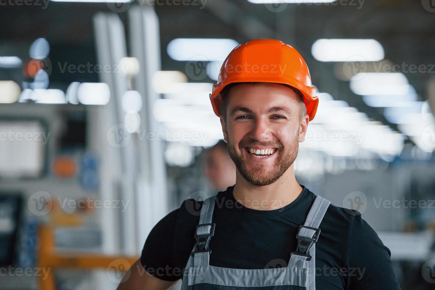 Smiling and happy employee. Portrait of industrial worker indoors in factory. Young technician with orange hard hat photo