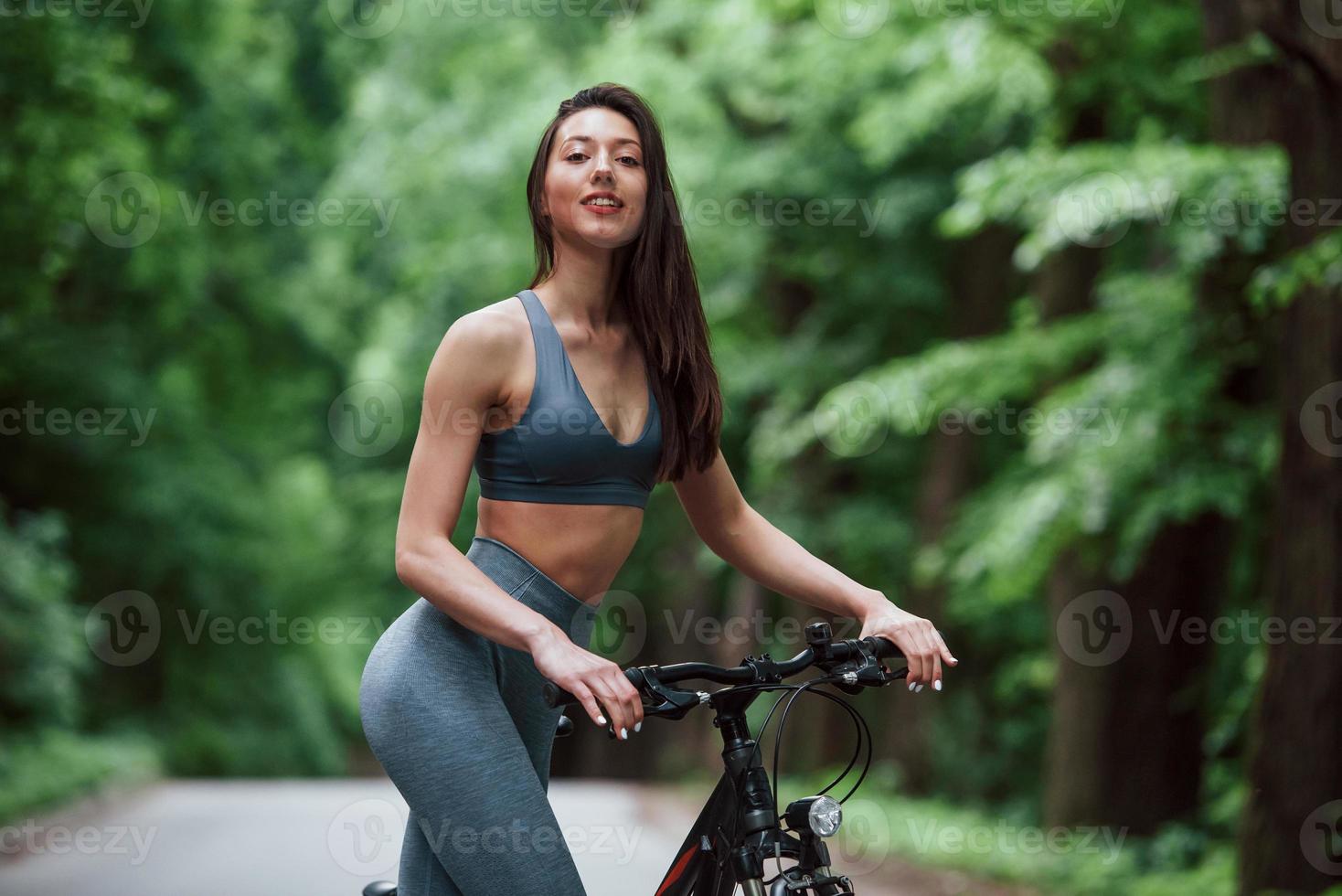 Positive emotions. Female cyclist standing with bike on asphalt road in the forest at daytime photo