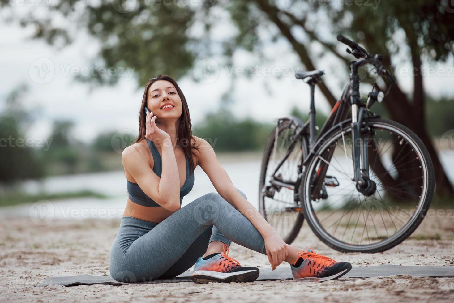 usando un teléfono inteligente. ciclista femenina con buena forma corporal sentada cerca de su bicicleta en la playa durante el día foto