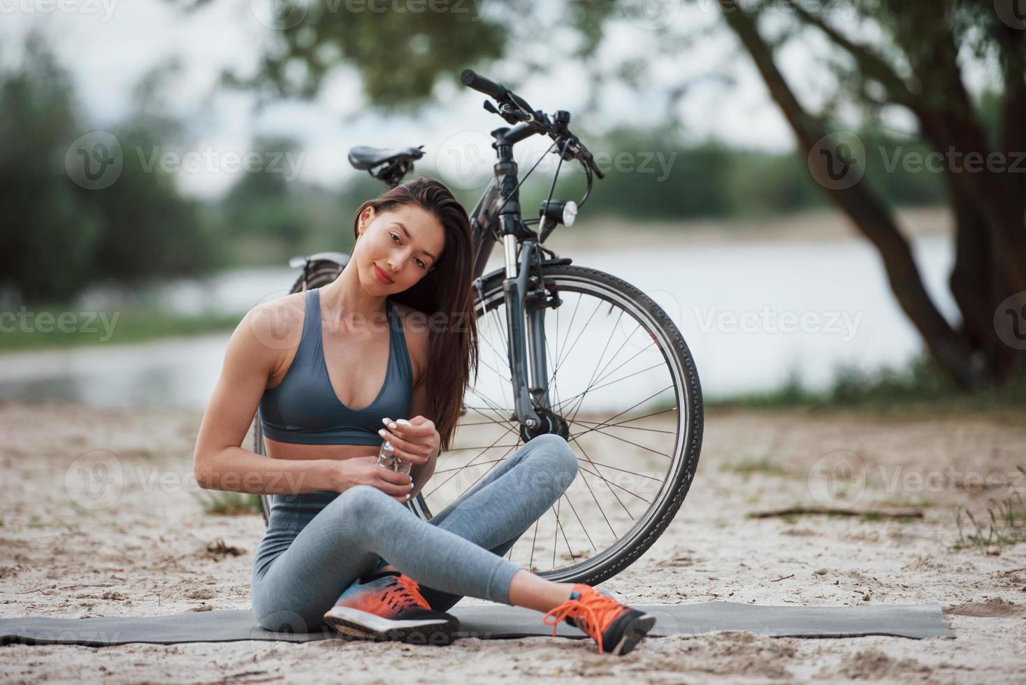 botella de apertura con agua. ciclista femenina con buena forma corporal sentada cerca de su bicicleta en la playa durante el día foto