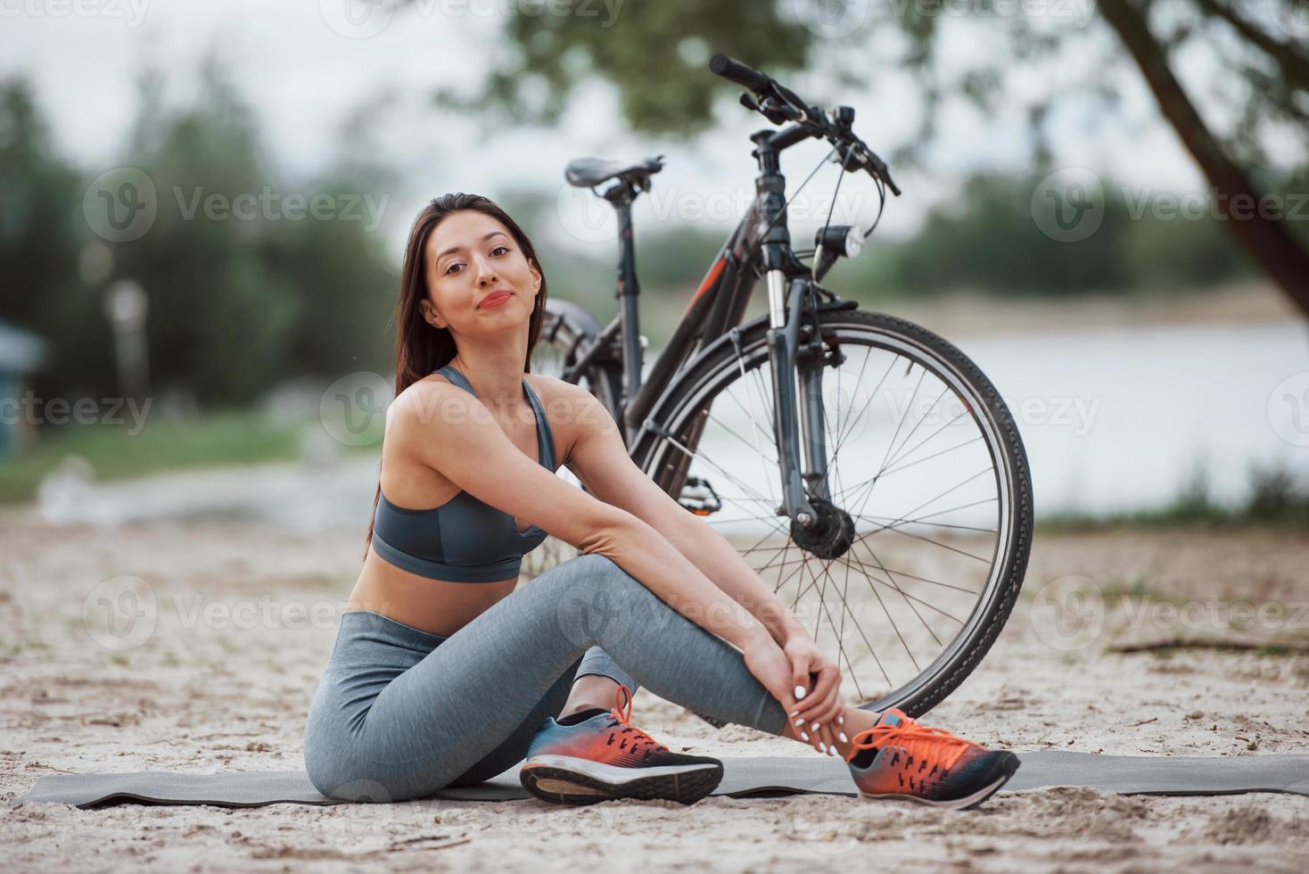 relajado y satisfecho. ciclista femenina con buena forma corporal sentada cerca de su bicicleta en la playa durante el día foto