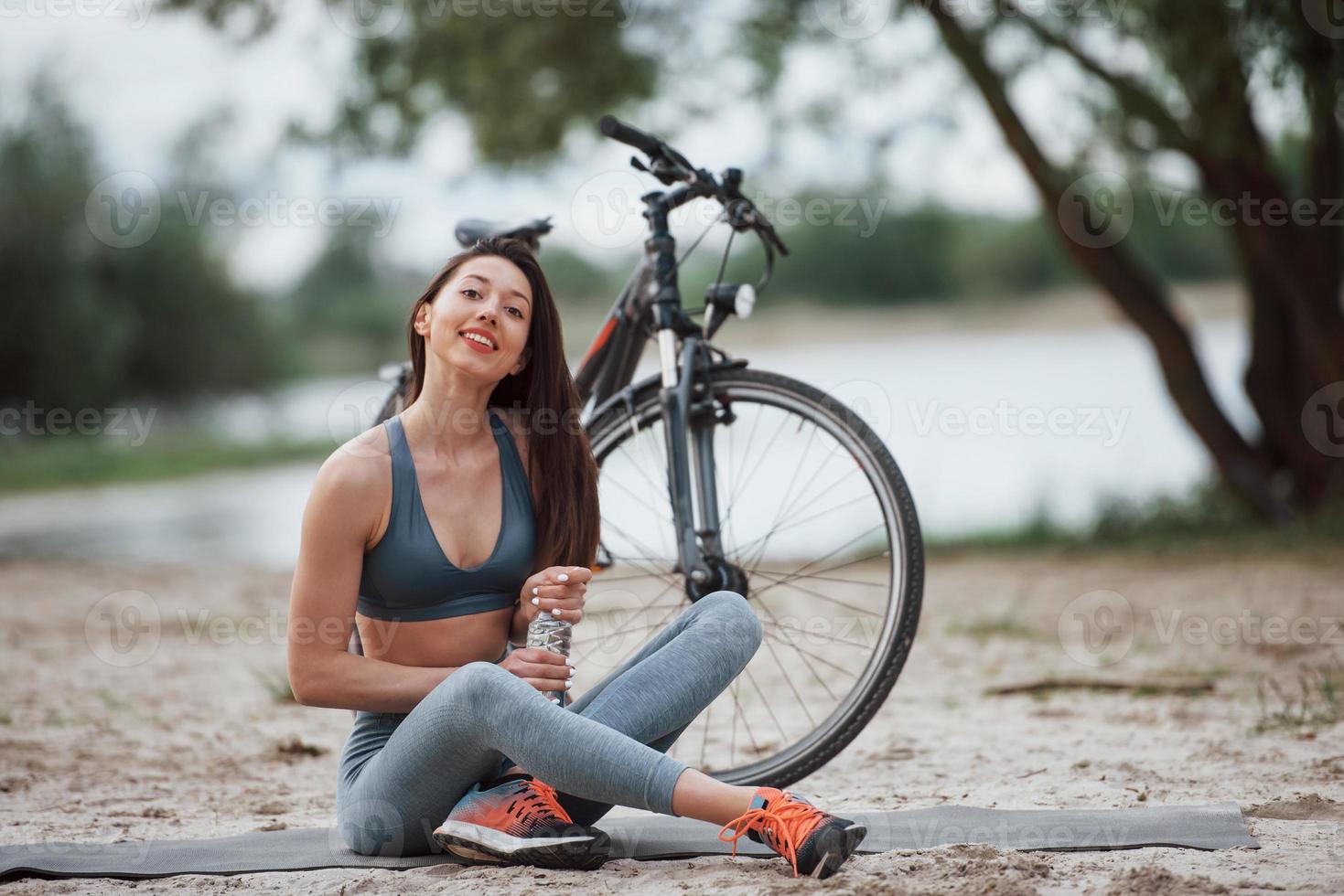 necesita recuperarse. ciclista femenina con buena forma corporal sentada cerca de su bicicleta en la playa durante el día foto