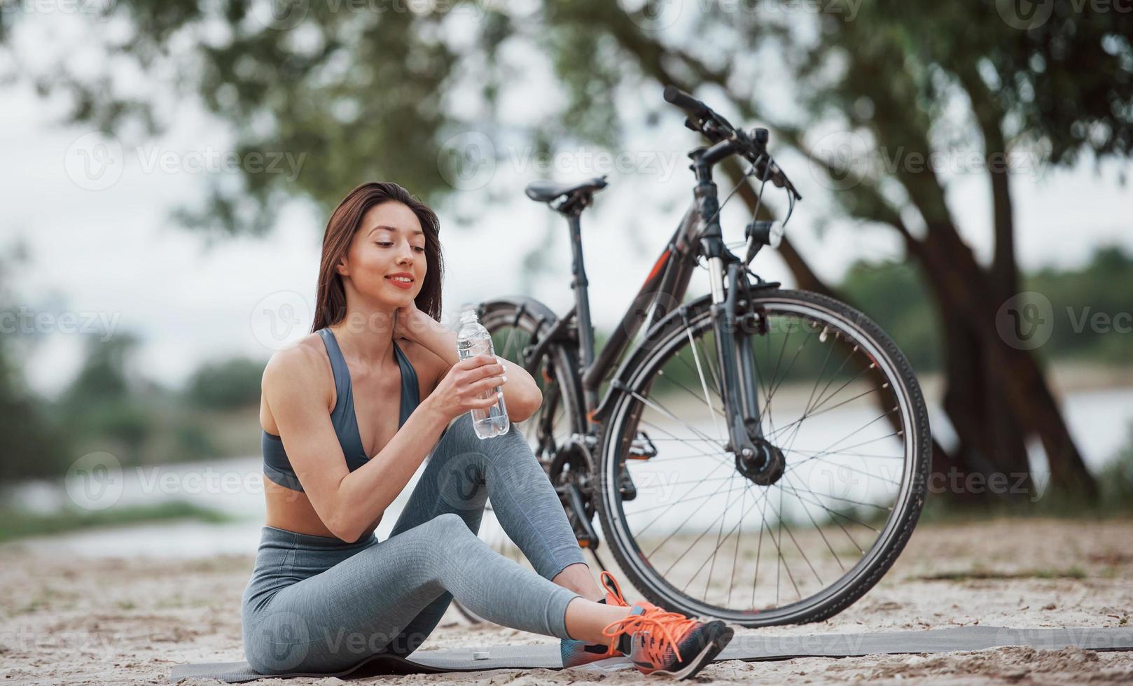 agua potable. ciclista femenina con buena forma corporal sentada cerca de su bicicleta en la playa durante el día foto