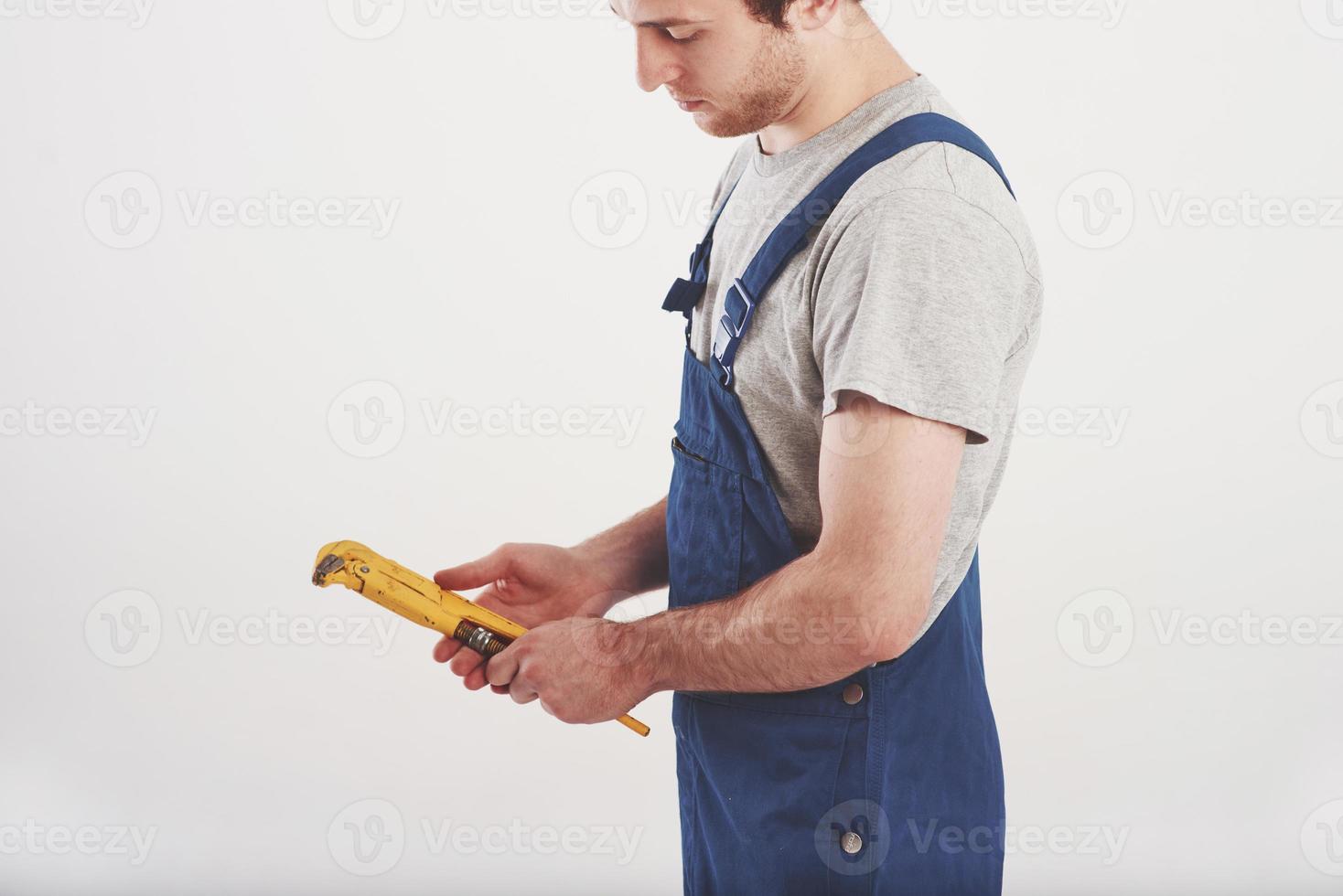 Looks at wrench. Man in blue uniform stands against white background in the studio photo