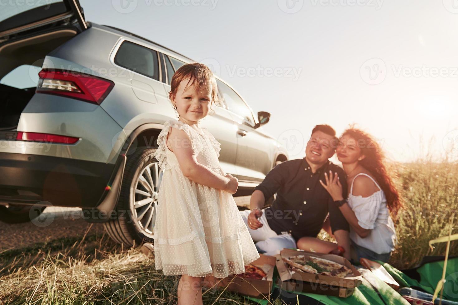 Cute dress on kid. Family have picnic at countryside near silver automobile at sunset photo