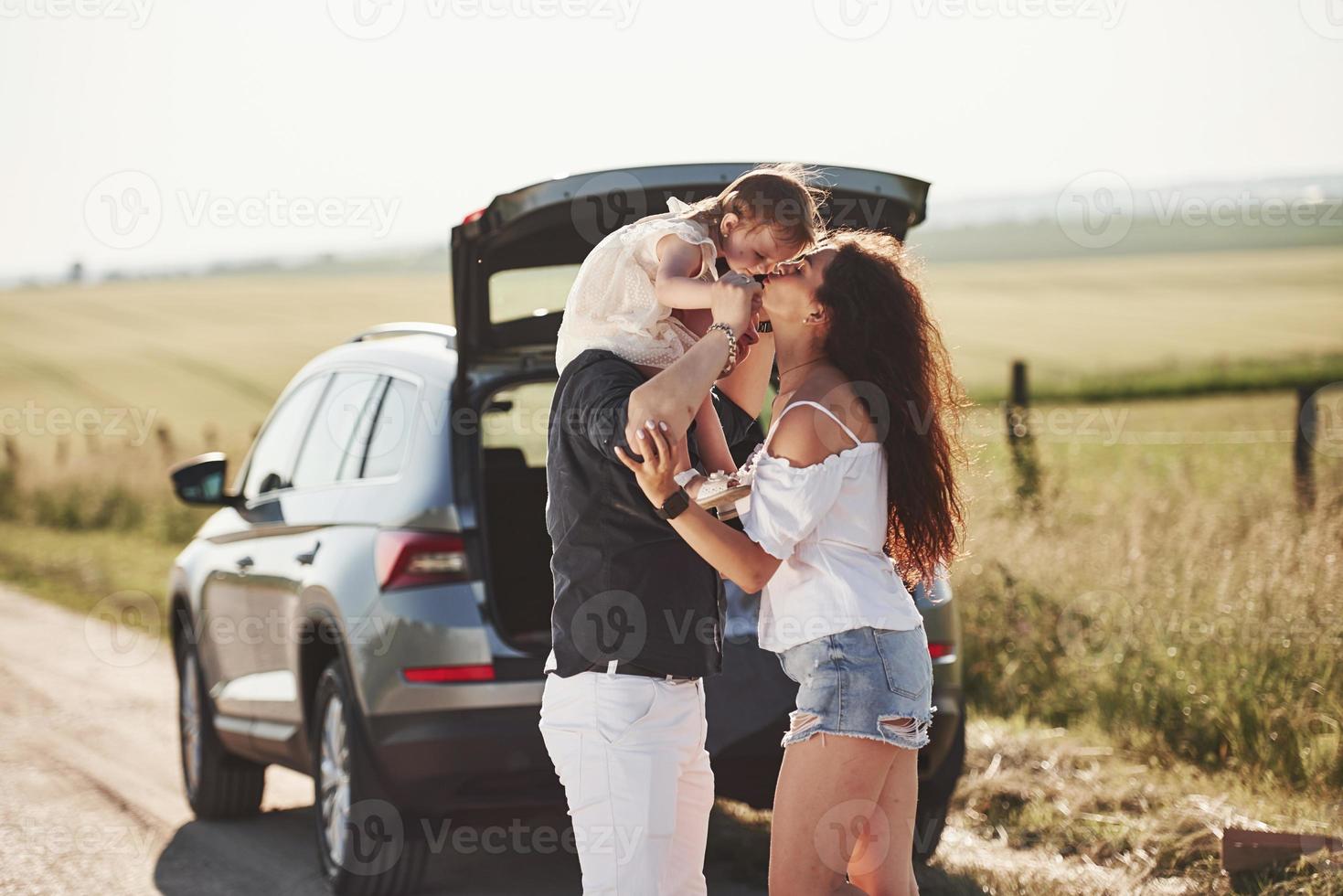 Mother kisses daughter. Family have some good time at countryside near silver automobile at sunset photo