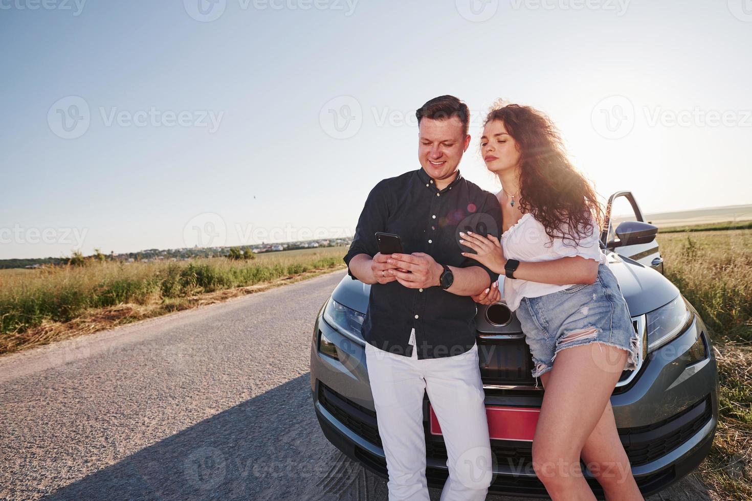 With phone in hands. Lovely couple near their new modern car at weekend time photo