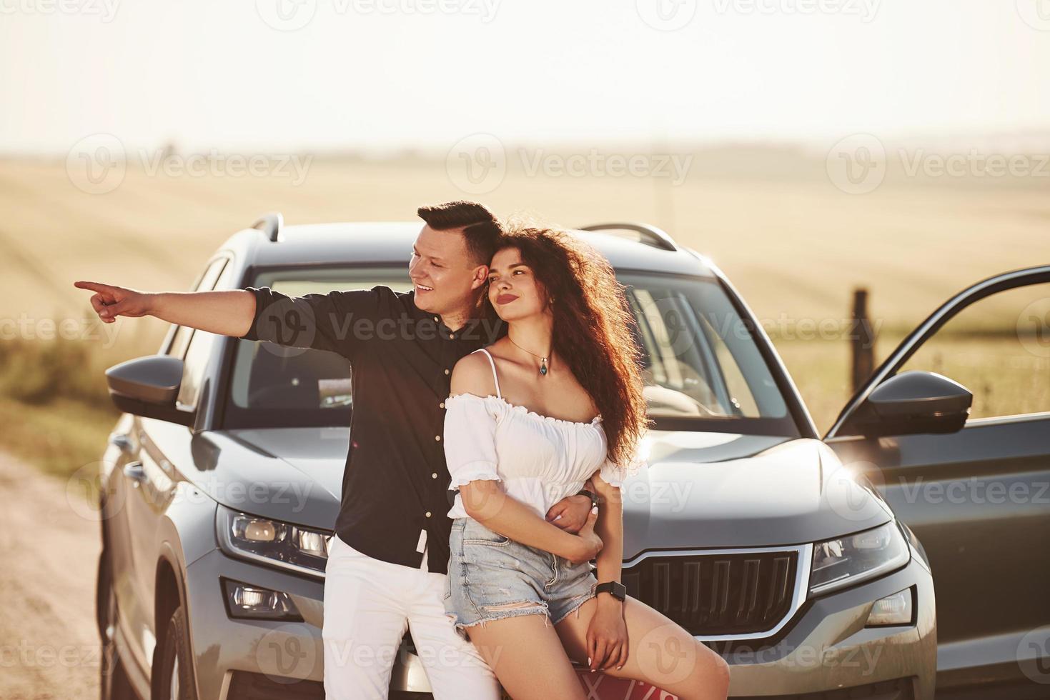 Shows something by the hand. Lovely couple near their new modern car at weekend time photo
