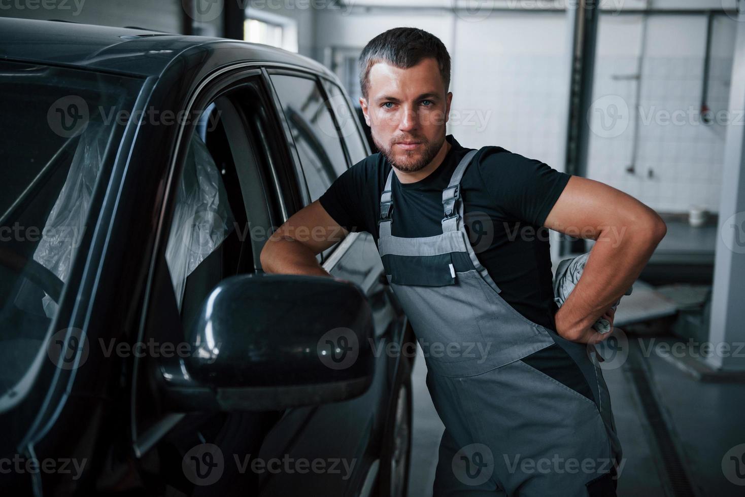 Looks straight at the camera. Young mechanic stands indoors near black car. After repairing automobile photo
