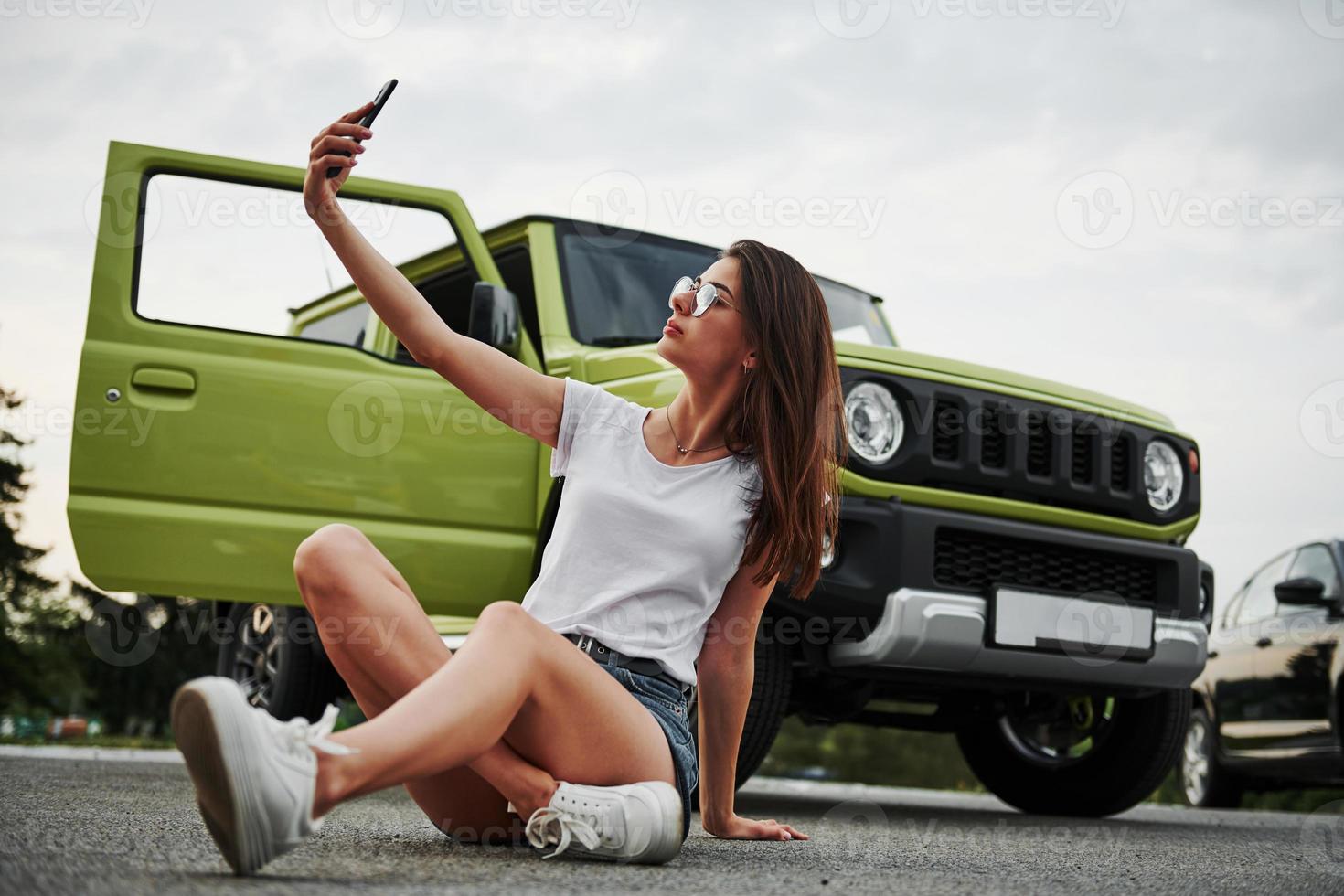 Making selfie. Sits and resting near new vehicle. Pretty woman in the green modern car posing for the camera photo