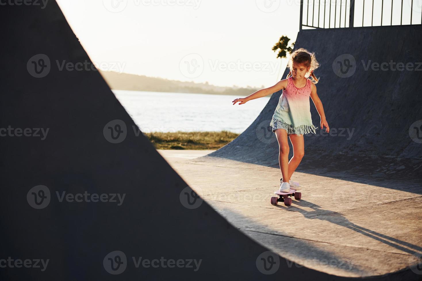 Sunny day. Kid have fun with skate at the ramp. Cheerful little girl photo