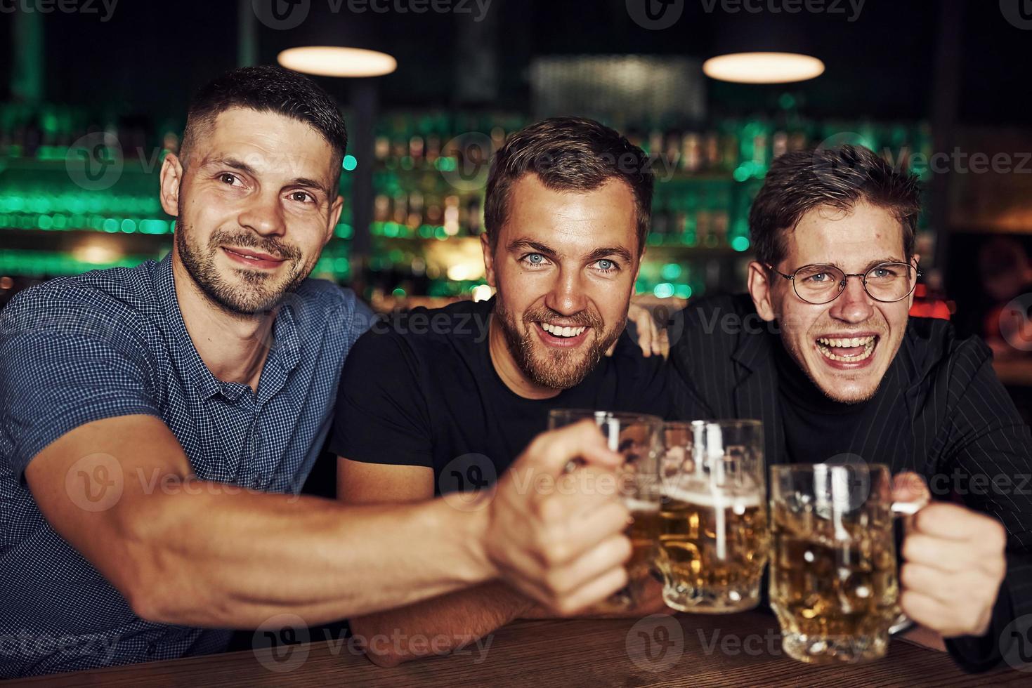 golpeando vasos. tres aficionados al deporte en un bar viendo fútbol. con cerveza en las manos foto